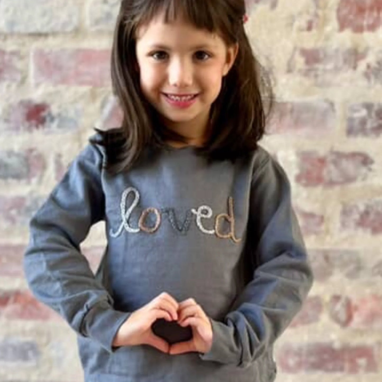 Girl wearing gray 'loved' sweatshirt, making heart shape with hands, brick wall background