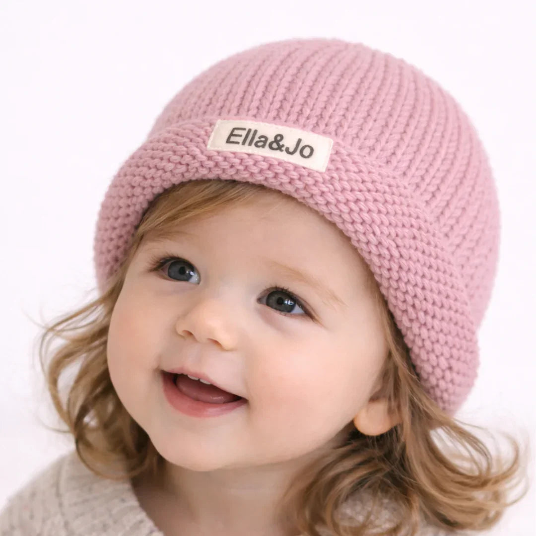 Smiling toddler wearing soft pink knit beanie with Ella&Jo label, cozy baby winter hat