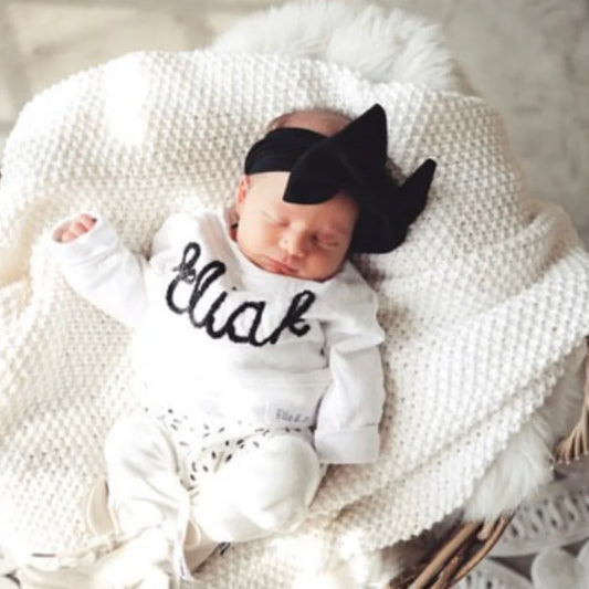Newborn baby in white 'Ella' onesie with black bow headband, cozy knit blanket, soft nursery setting