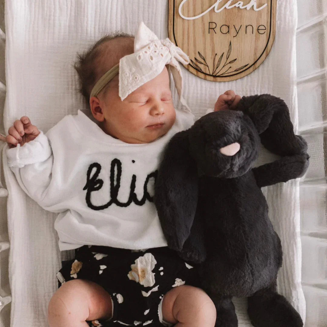 Newborn baby in white personalized top and floral shorts, cuddling black bunny plush toy