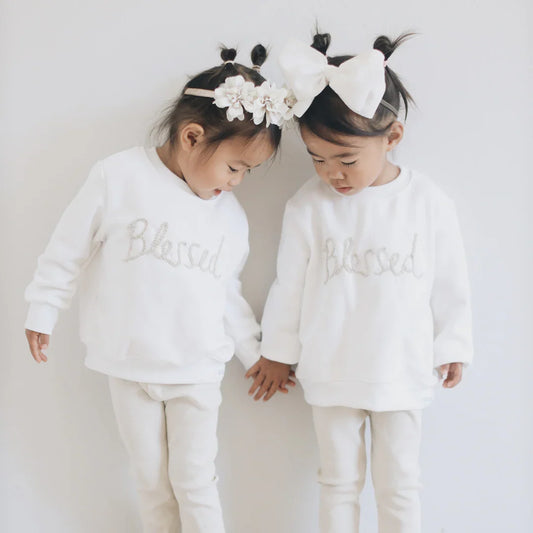 Two little girls in matching white 'Blessed' sweaters and pants, holding hands with floral headbands.