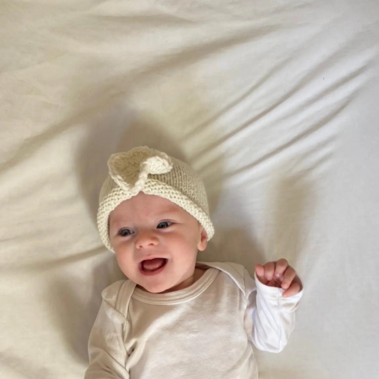 Baby wearing cream knit hat with bow, smiling on soft blanket