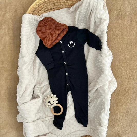 Black baby onesie with smiley face, brown knit hat, and lion teether on white minky blanket