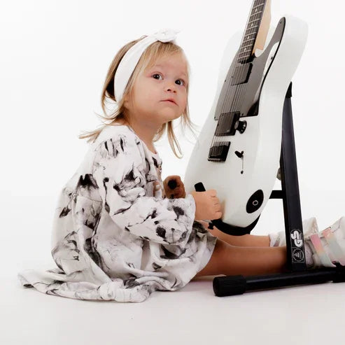 Toddler girl in black and white tie-dye dress holding white electric guitar on stand