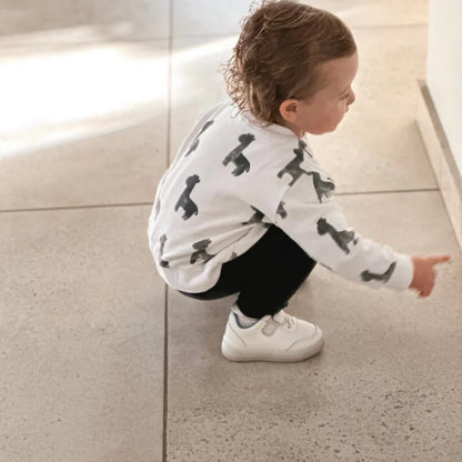 Toddler in white dino-print sweater, black pants, and white sneakers pointing on tiled floor