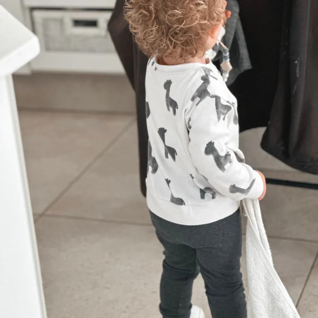 Toddler in white dinosaur print sweatshirt and dark grey pants, holding a towel