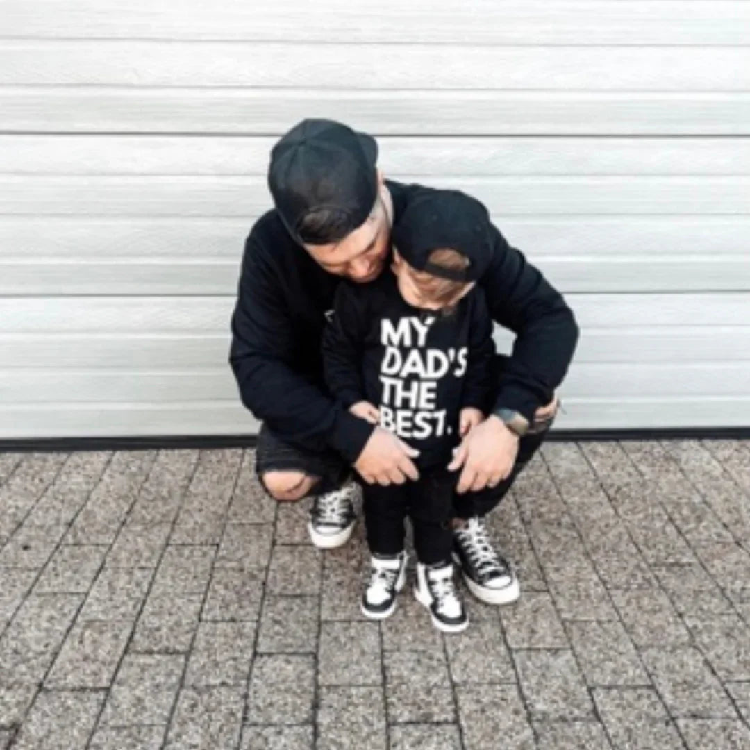 Dad and toddler in matching black outfits, child's shirt says 'MY DAD'S THE BEST', casual family style