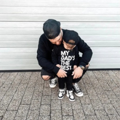 Dad and toddler in matching black outfits, child's shirt says 'MY DAD'S THE BEST', casual family style
