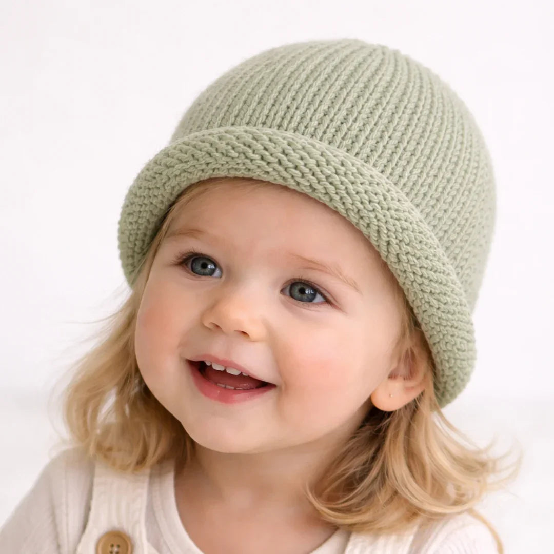 Smiling toddler wearing soft sage green knit beanie hat, cozy baby clothing